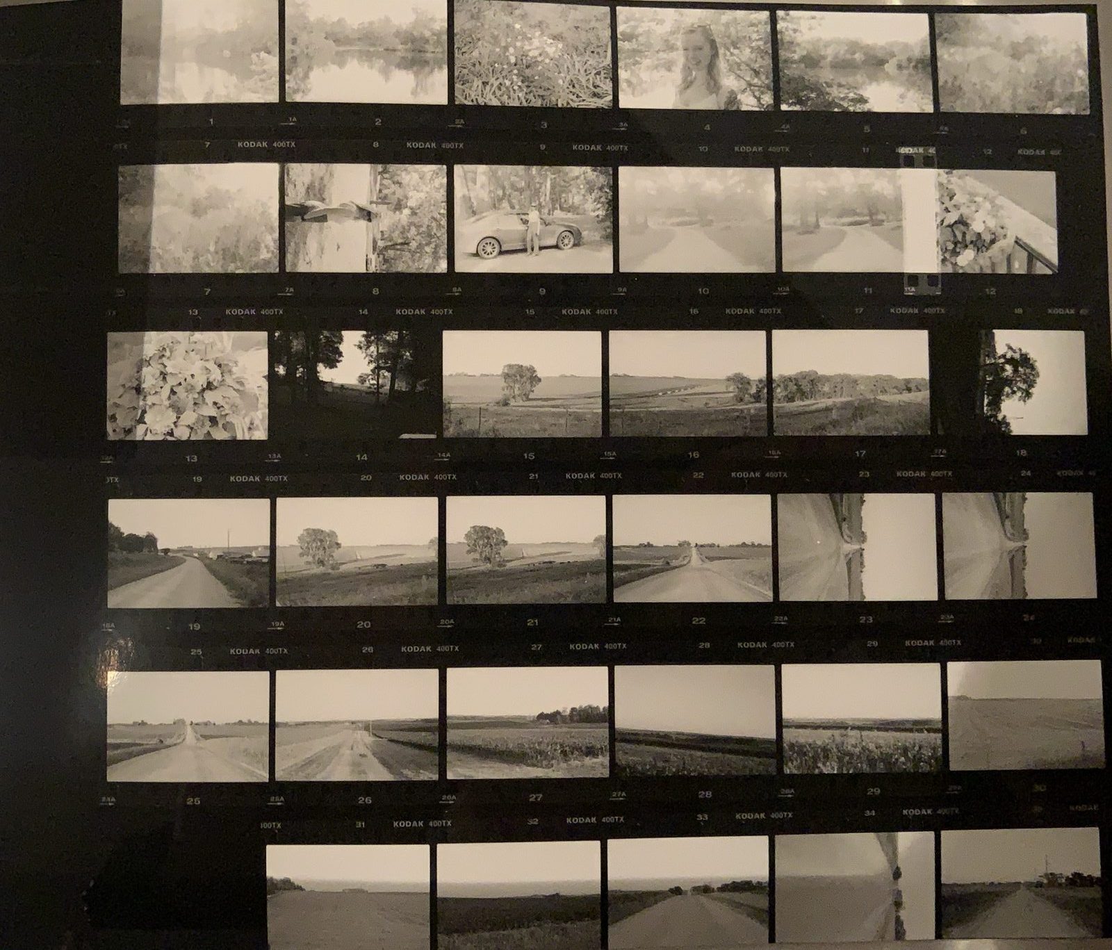 Black-and-white contact sheet of thirty-some frames showing roads, fields, a car, and flowers