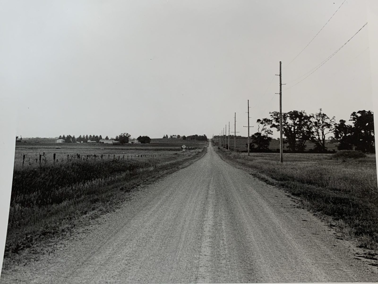 Black-and-white print of a gravel road running to the horizon beside a line of utility poles