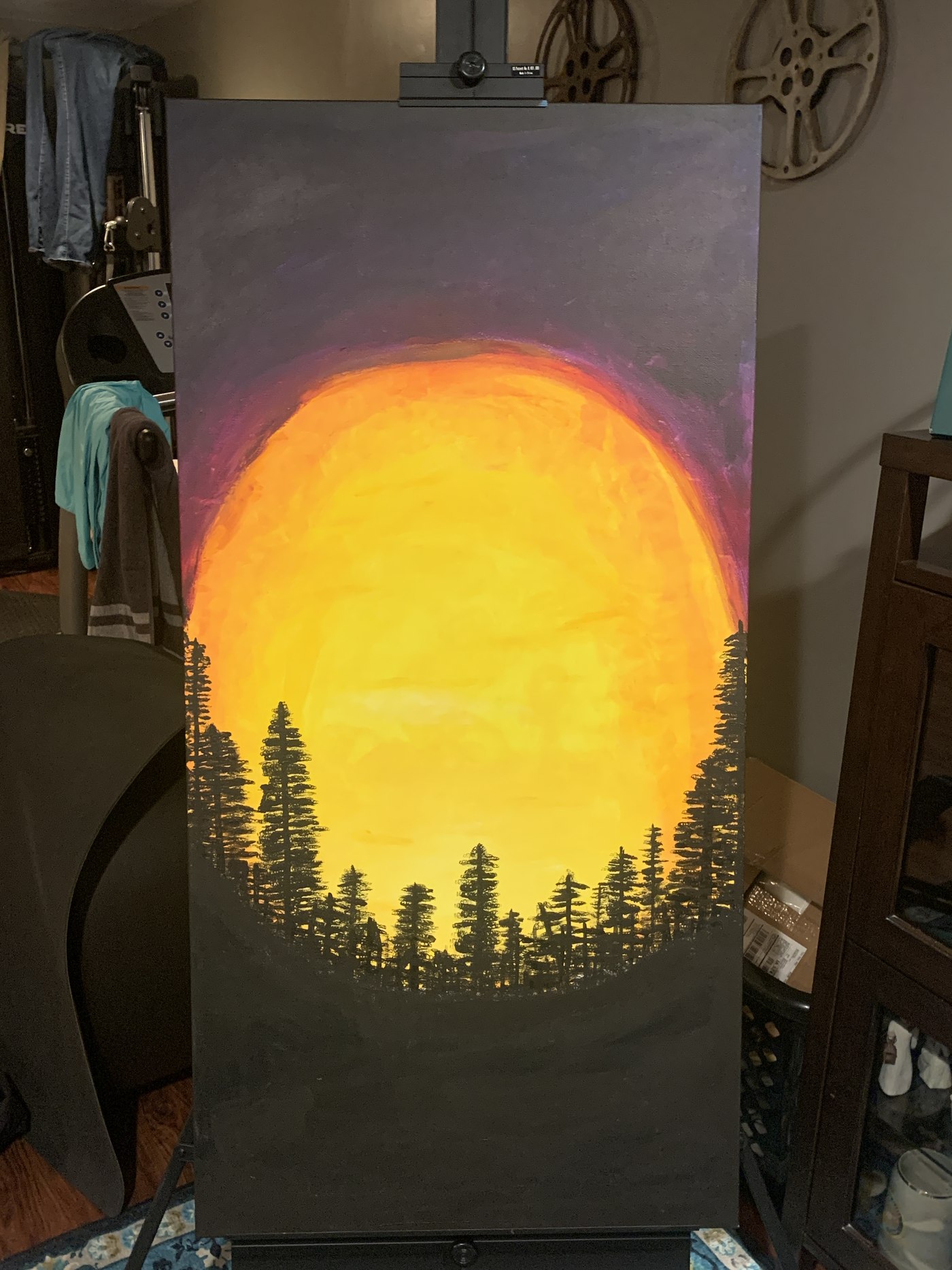 Acrylic painting of an orange sun setting behind a silhouette of pine trees