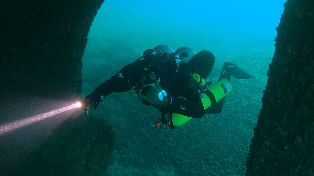 A diver with a green stage bottle swimming through a wreck opening